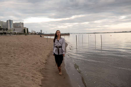 A beautiful young brunette woman walks on the beach in cool weather in a fashionable outfitの写真素材