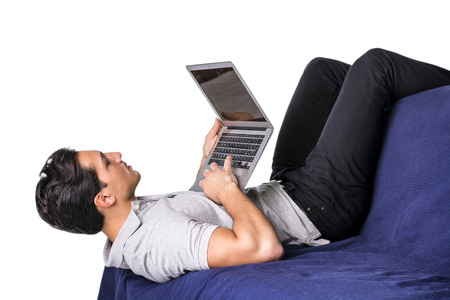 Young attractive man working with computer while laying in awkward position on couch, on white backgroundの写真素材