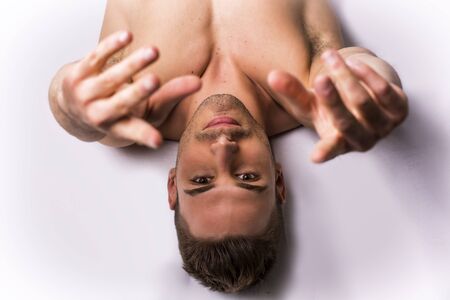 Shirtless young man leaning on white sheet, shot from above. Hands over his head, looking at camera. Muscular buildの写真素材