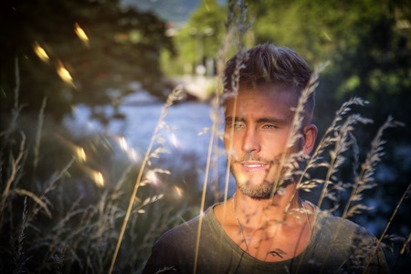 Blondish, blue eyed young man by river at sunset, thinking, looking awayの写真素材