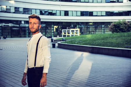 Blond Young Man Next to Modern Building in City, Wearing White Shirt and Suspenders, Standingの写真素材