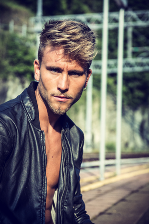 Attractive blond young man standing on railroad tracks, wearing only black leather jacket, looking at cameraの写真素材