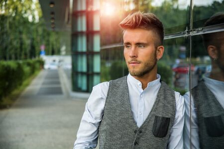 Blond Young Man Next to Modern Building in City, Wearing White Shirt and Elegant Vest, Standing and Leaning against Glass Wallの写真素材
