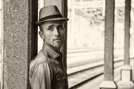 Portrait of blond haired handsome young man with fedora hat. An old train station is behind him in...