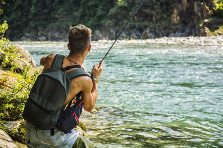Young handsome male tourist standing and fishing in river in sunny day with rodの写真素材