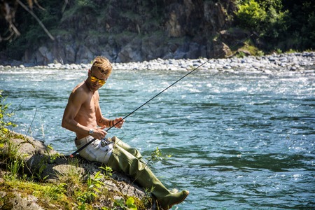 Young handsome male tourist standing and fishing in river in sunny day with rodの写真素材