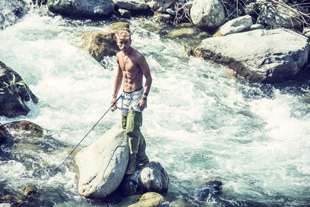 Young handsome male tourist standing and fishing in river in sunny day with rodの写真素材