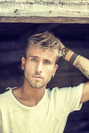 Handsome blond, blue eyed young man leaning against wood planks wall outdoor and looking away to a sideの写真素材