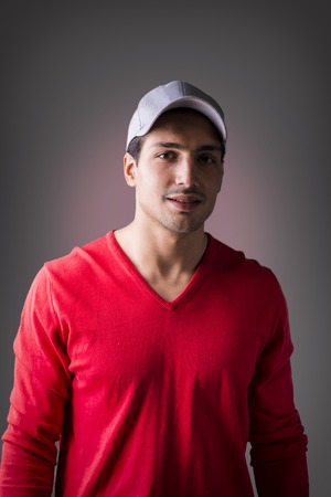 A young man with red wool sweater, wearing baseball cap, front view. On dark backgroundの写真素材