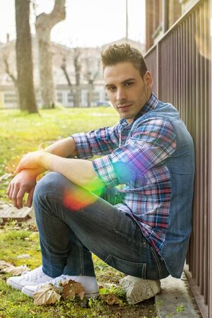 An attractive young man sitting on ground outdoor in public park against metal fence, wearing jeans and shirtの写真素材
