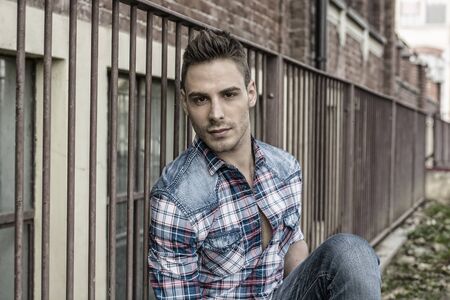 An attractive young man sitting on ground outdoor in public park against metal fence, wearing jeans and shirtの写真素材