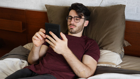 Handsome young man at home reading with ebook reader lying on his bed