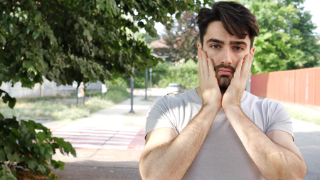 Young exhausted man holding hands on cheeks looking unhappily at camera on background of street.の写真素材