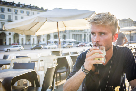 Handsome blonde young man sitting in Turin cafe holding glass wearing black t-shirt looking away.の写真素材