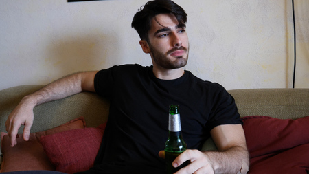 Young attractive man sitting on couch at home in living-room, drinking beer, relaxing in a lazy...