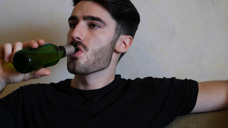 Young attractive man sitting on couch at home in living-room, drinking beer, relaxing in a lazy afternoonの写真素材