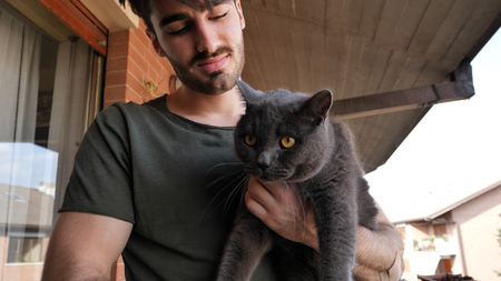 Handsome Young Animal-Lover Man Outside the House, Holding, Hugging and Petting his Gray Domestic Cat Pet.の写真素材