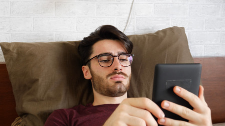 Young handsome man lying in bed and reading an ebook in his bedroom at home at night