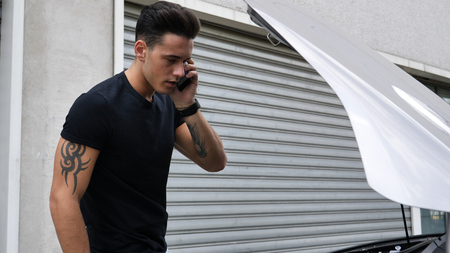 Young man trying to repair a car engine and seeking help with cell phone calling a mechanic