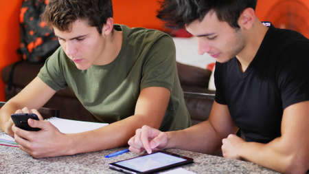 Two casual male friends posing at table with devices and notepads studying together, doing homework