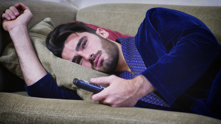Young bored sleepy man sitting on sofa watching television changing the channel with the remote...