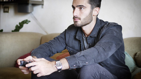 Attractive young man using joystick or joypad for videogames, sitting on couch at home in his living...
