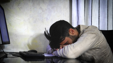 Young man sleeping on desk in front of computer at home, tired from working late at nightの写真素材
