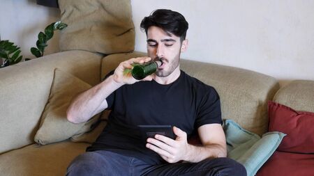 Young attractive man sitting on couch at home in living-room, drinking beer and reading book from ebook reader, relaxing in a lazy afternoonの写真素材