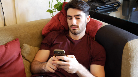 Young man typing and using a cell phone while sitting at home on a couch to send messages or surf...