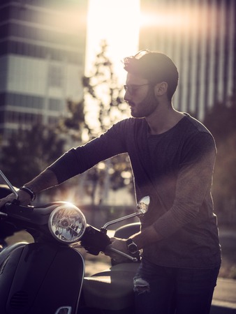 Young handsome man sitting on scooter in backlit on city street.