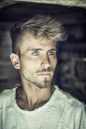 Handsome blond, blue eyed young man leaning against wood planks wall outdoor and looking away to a...
