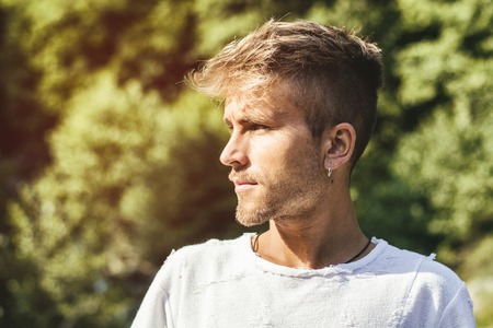 Sad, worried blond young man against trees, outside in nature, looking to a sideの写真素材