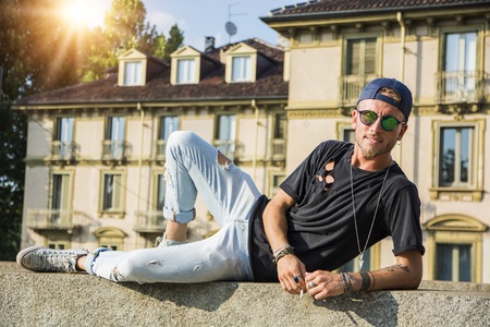 Young trendy hipster man in casual clothing posing on stone fence at street in sunlight looking at camera smilingの写真素材