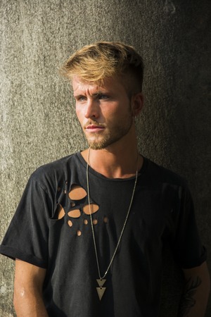 Handsome blond, blue eyed young man leaning against stone wall outdoor and looking to a side in Turin, Italyの写真素材