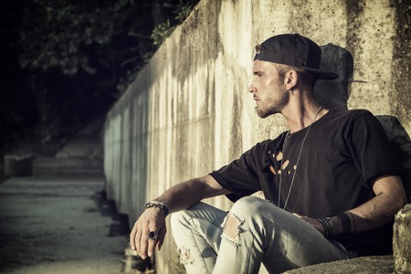 Handsome blond, blue eyed young man leaning against stone wall outdoor and looking to a sideの写真素材