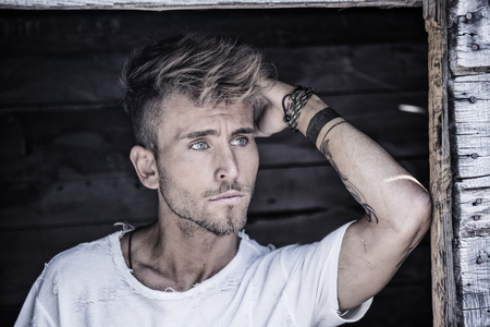 Handsome blond, blue eyed young man leaning against wood planks wall outdoor and looking away to a sideの写真素材