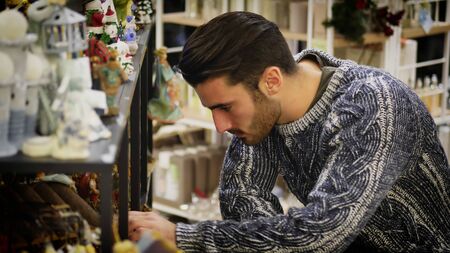Handsome young man in jumper standing near shelf in shop and looking at goods.の写真素材