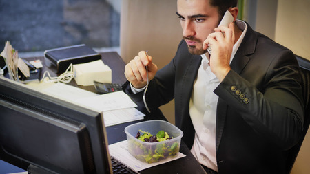 Elegant man in suit having lunch at table in office while talking on phone and watching computer.の写真素材