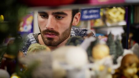 Handsome young man in jumper standing near shelf in shop and looking at goods.