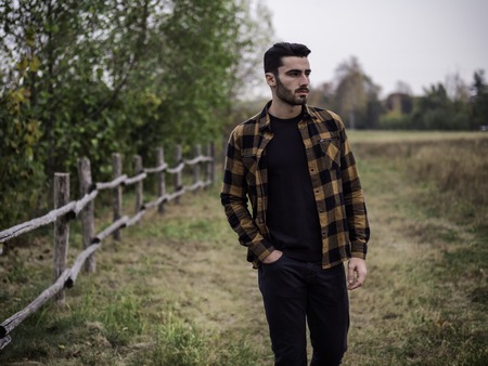 Portrait of sexy young man looking to a side near wooden fence in countryside next to a field or...