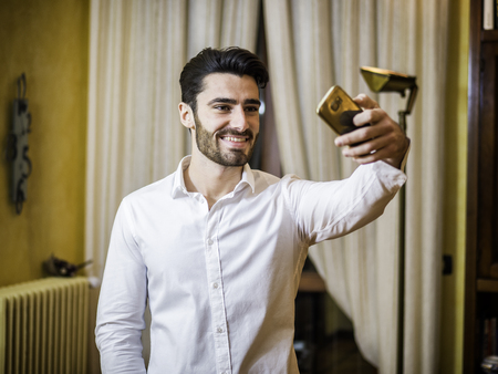 Handsome smiling young man doing selfie with smartphone at home in living room. Horizontal indoors shotの写真素材