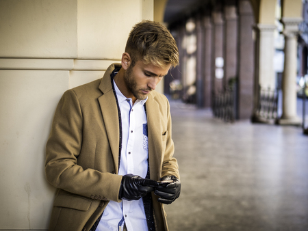 Handsome trendy blond man wearing dark coat standing and looking down at a cell phone that he is holding, outdoor in European city setting with elegant old historic building behindの写真素材