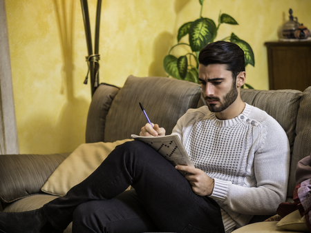 Young man sitting doing a crossword puzzle looking thoughtfully at a magazine, with his pencil to his mouth, as he tries to think of the answer to the clueの写真素材
