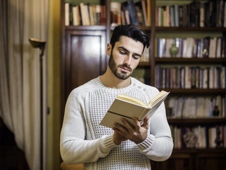 Handsome young man reading book at home in his living-room, standingの写真素材