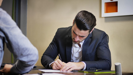 Successful handsome young businessman sitting at his desk in the office and signing document on...