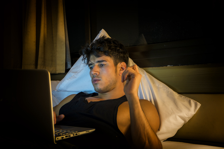 Attractive Young Man with Serious Expression, Reclining Comfortably with Laptop on Bed Working on his Start-up Business - Young Male College or University Student Doing Homework, in Bedroomの写真素材