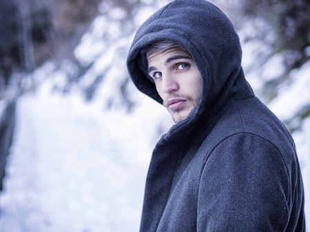 Portrait of young man in hoodie posing outdoor in winter setting with snow all around, looking at...