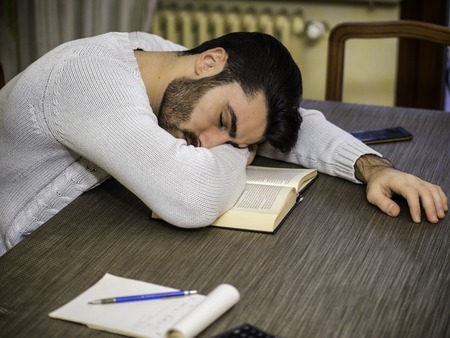 Over-worked, tired young man at home sleeping instead of working or studying, resting head over book. Tired male student falling asleep reading bookの写真素材