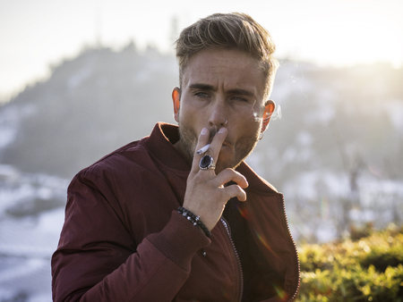 Handsome young man outdoor in winter fashion smoking cigarette, wearing black coat and woolen scarf in city parkの写真素材