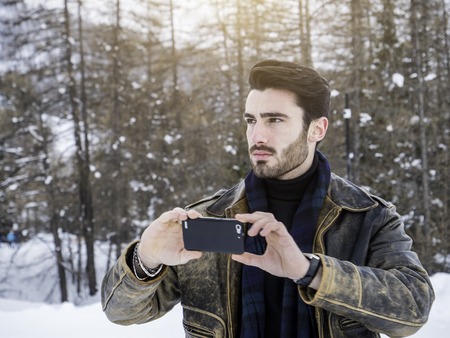 Handsome young man using smarpthone to take photographs of the landscape, while posing among snowy...
